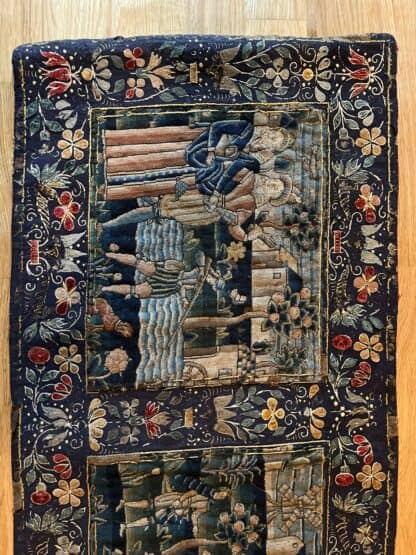 left most part of the needlework panel, with the first scene in the centre showing a couple of parents looking on at their son with a rifle